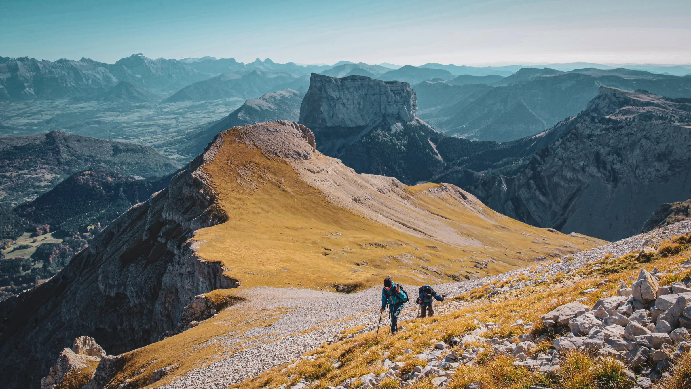 Two hikers climb a golden ridge with majestic mountains in the background.