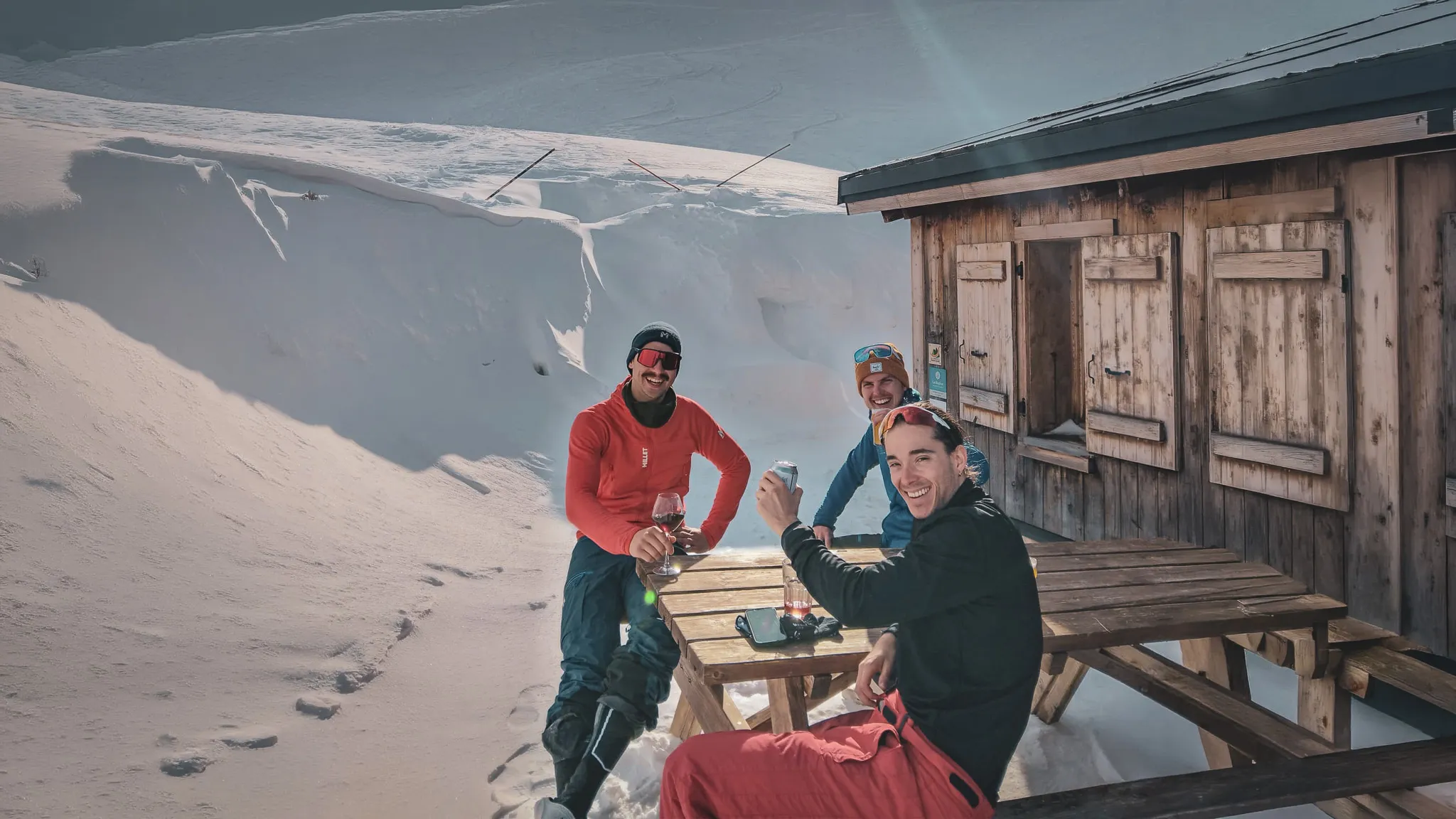Drie vrienden genieten van de zon en een drankje voor een berghut, omringd door sneeuw.