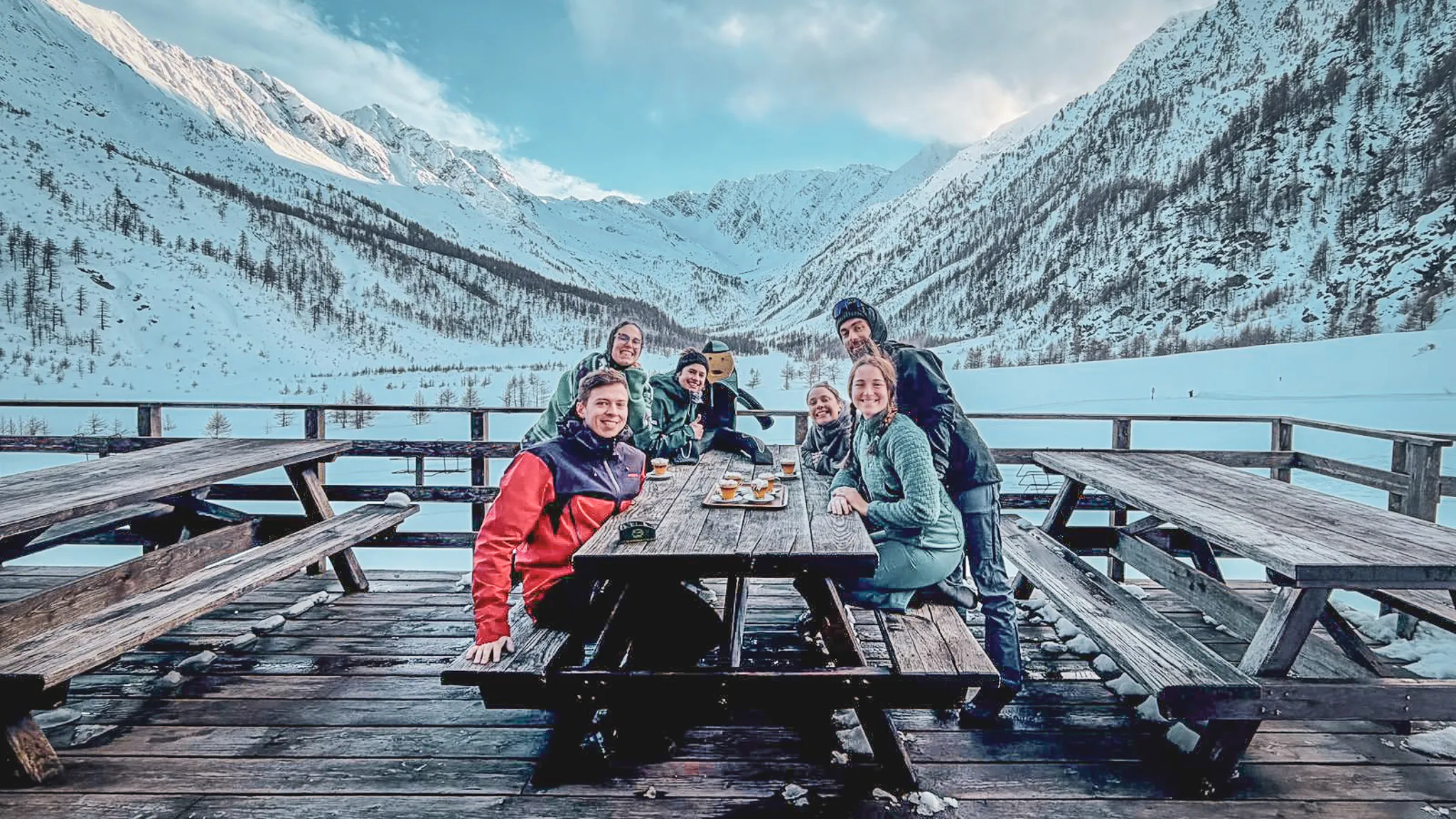Groep van zes lachende deelnemers op een houten terras met besneeuwde bergen op de achtergrond