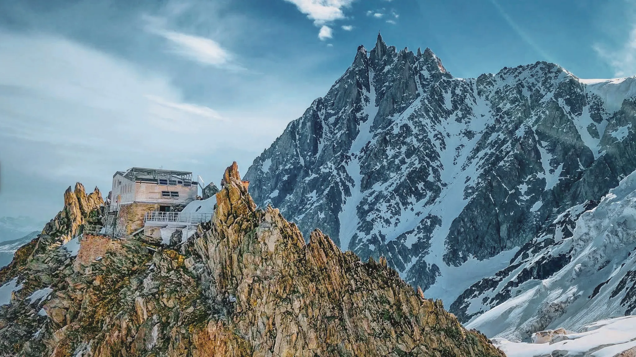 A mountain hut perched on the rocks, with snow-capped peaks in the background.