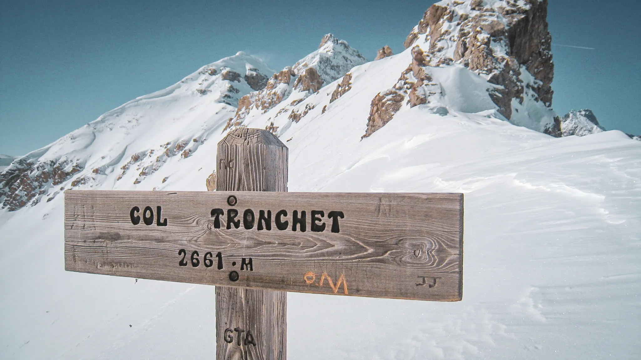 Wooden sign indicating the Col Tronchet, with snow-capped mountains in the background.