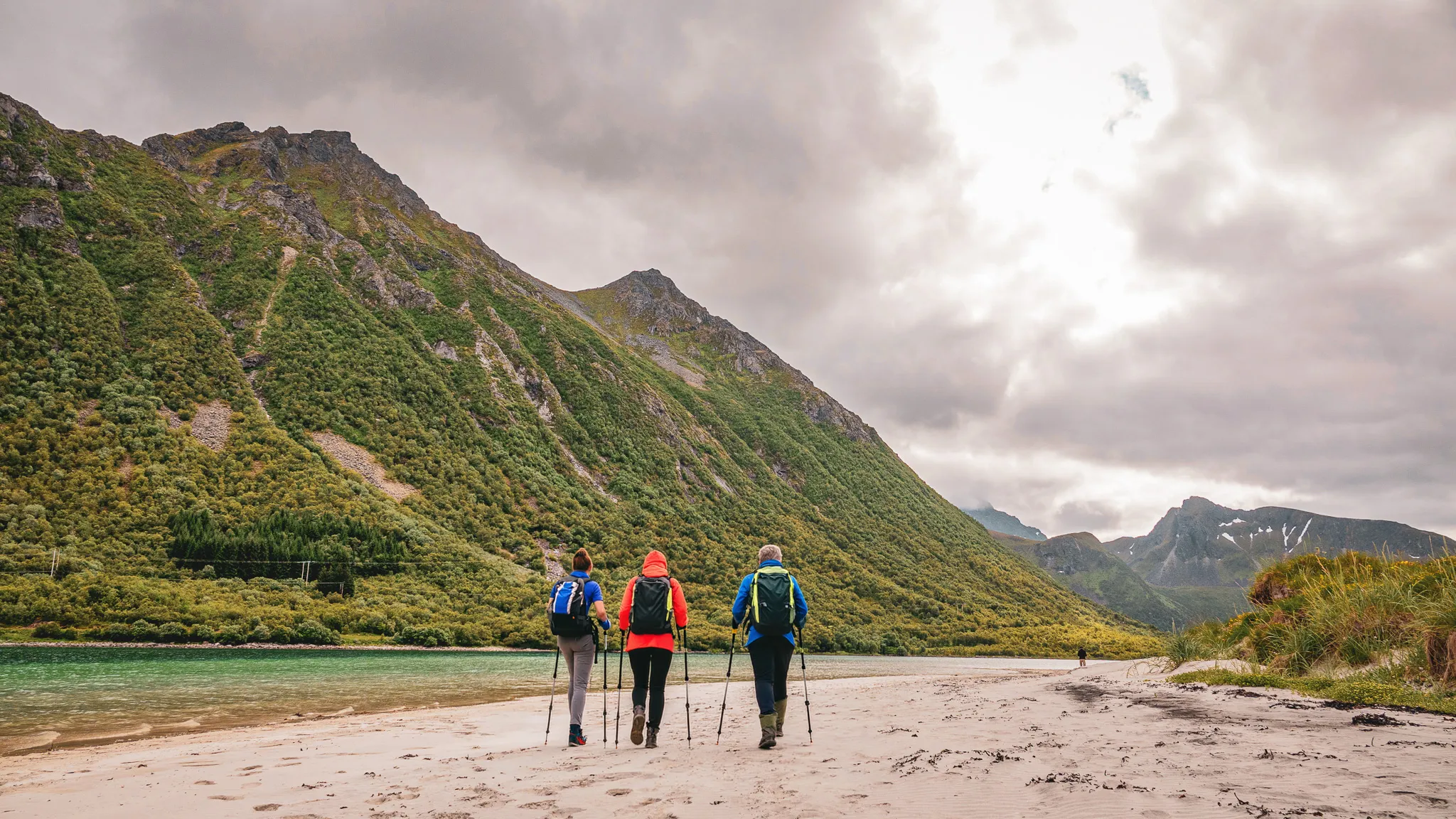 Three hikers walking on a beach, surrounded by green mountains in the Lofoten Islands.