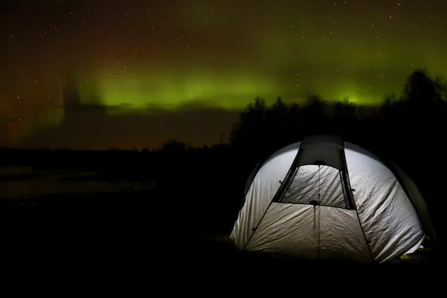 photo de nuit du Kungsleden, où l'on voit des aurores boréales verte dans un ciel étoilé, avec en premier plan une tente grise éclairer à l'intérieur 