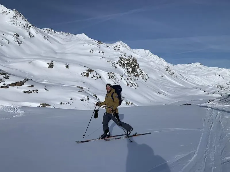 un homme pratiquant le ski de randonnée vétu de skis, chaussures de skis, de batons de skis, d'un pantalon gris, d'une veste marron, des gants noir et un sac à dos noir. on peut le voir sur une montagne créant ses première traces et en fond des pics de montagnes et un ciel bleu. 